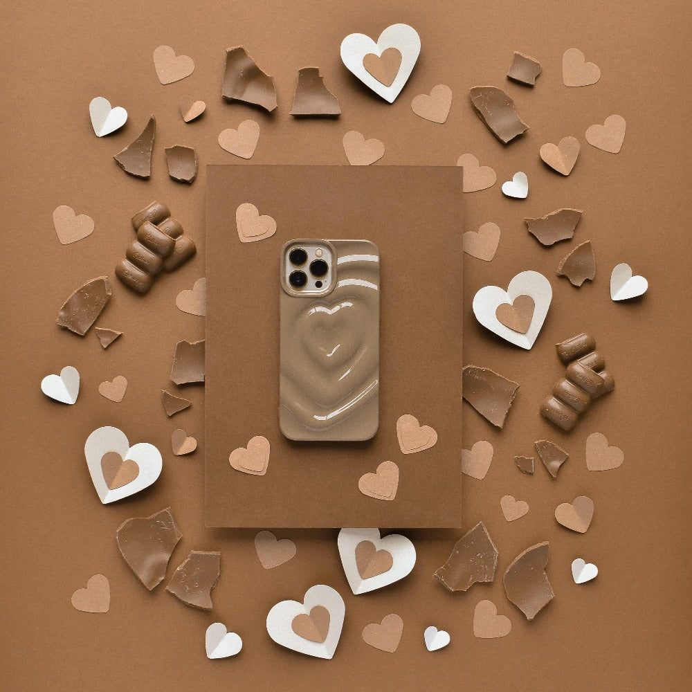 A Coconut Lane Melting Heart Phone Case in chocolate brown, featuring a beige and white wavy heart pattern, rests on a brown surface amidst heart-shaped paper cutouts and pieces of chocolate. The harmonious shades of chocolate brown in the background, chocolates, and decorations beautifully accentuate the elegant design of the phone case.