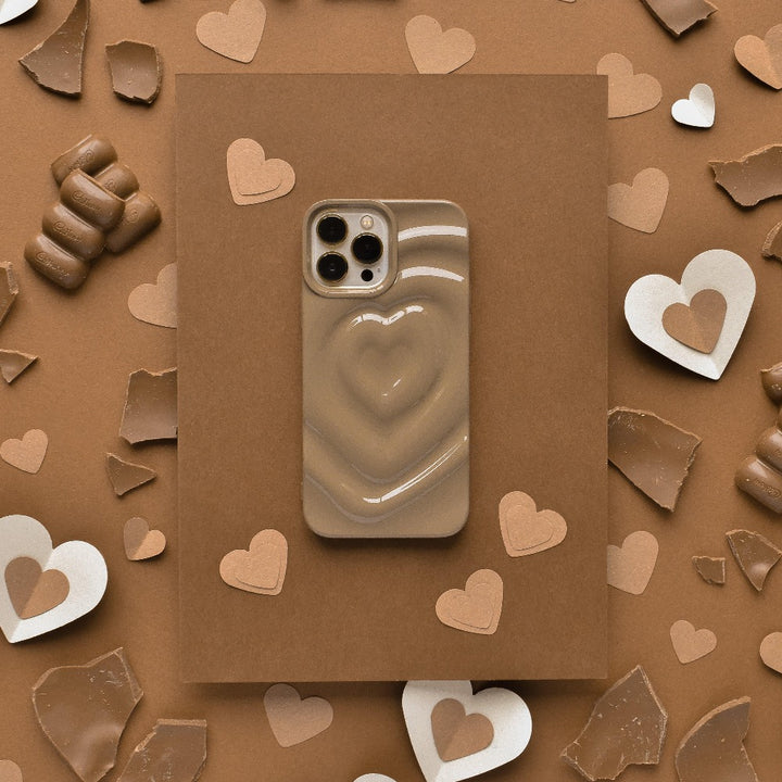 A Coconut Lane Melting Heart Phone Case in Chocolate Brown is placed on a brown background, surrounded by broken pieces of chocolate and scattered paper hearts. The color scheme predominantly features shades of chocolate brown and beige, creating a warm, cohesive look.