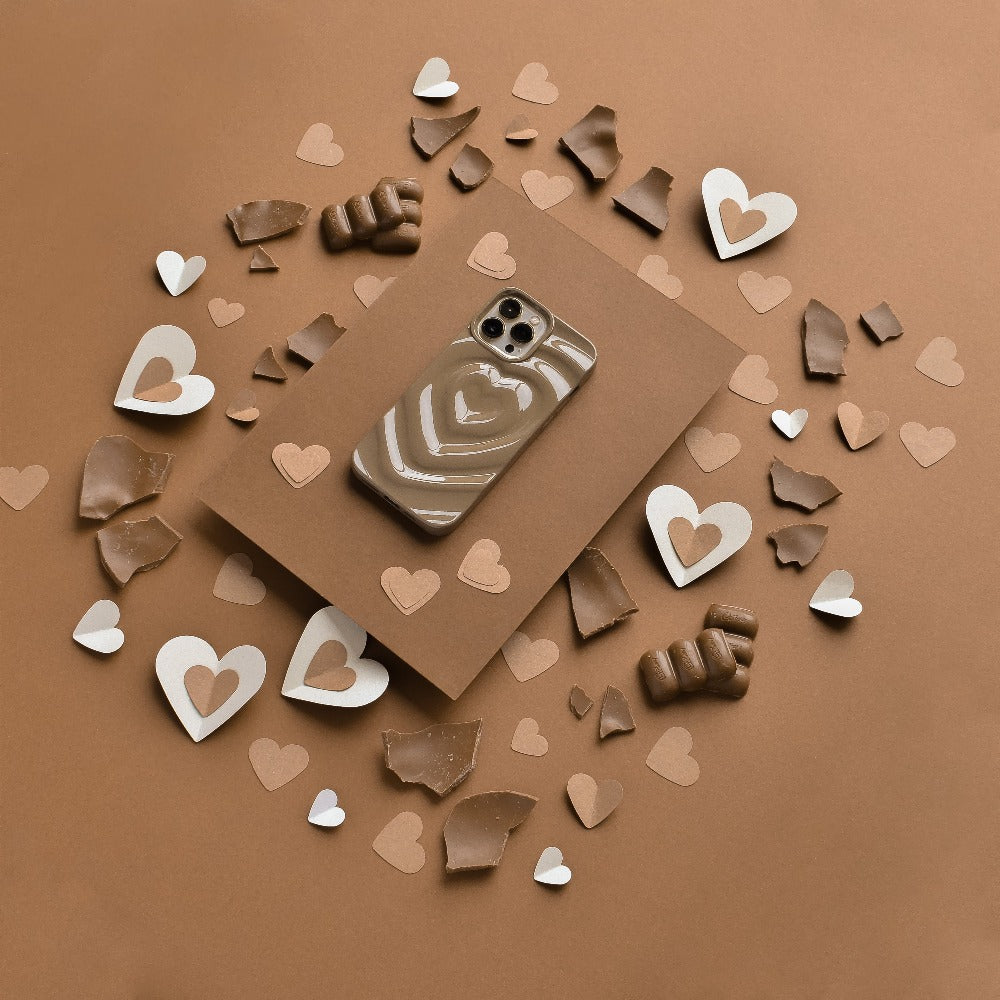 A smartphone encased in a Coconut Lane Melting Heart Phone Case - Chocolate Brown rests on a brown envelope. Surrounding the envelope are scattered pieces of brown and white paper and chocolate molded into various heart shapes and fragments, all set against a brown background.