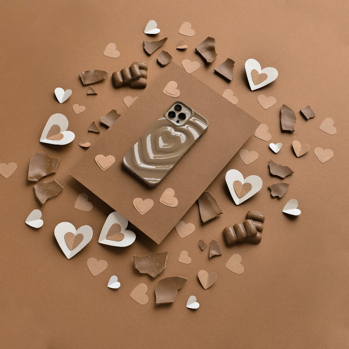 A smartphone encased in a Coconut Lane Melting Heart Phone Case - Chocolate Brown rests on a brown envelope. Surrounding the envelope are scattered pieces of brown and white paper and chocolate molded into various heart shapes and fragments, all set against a brown background.