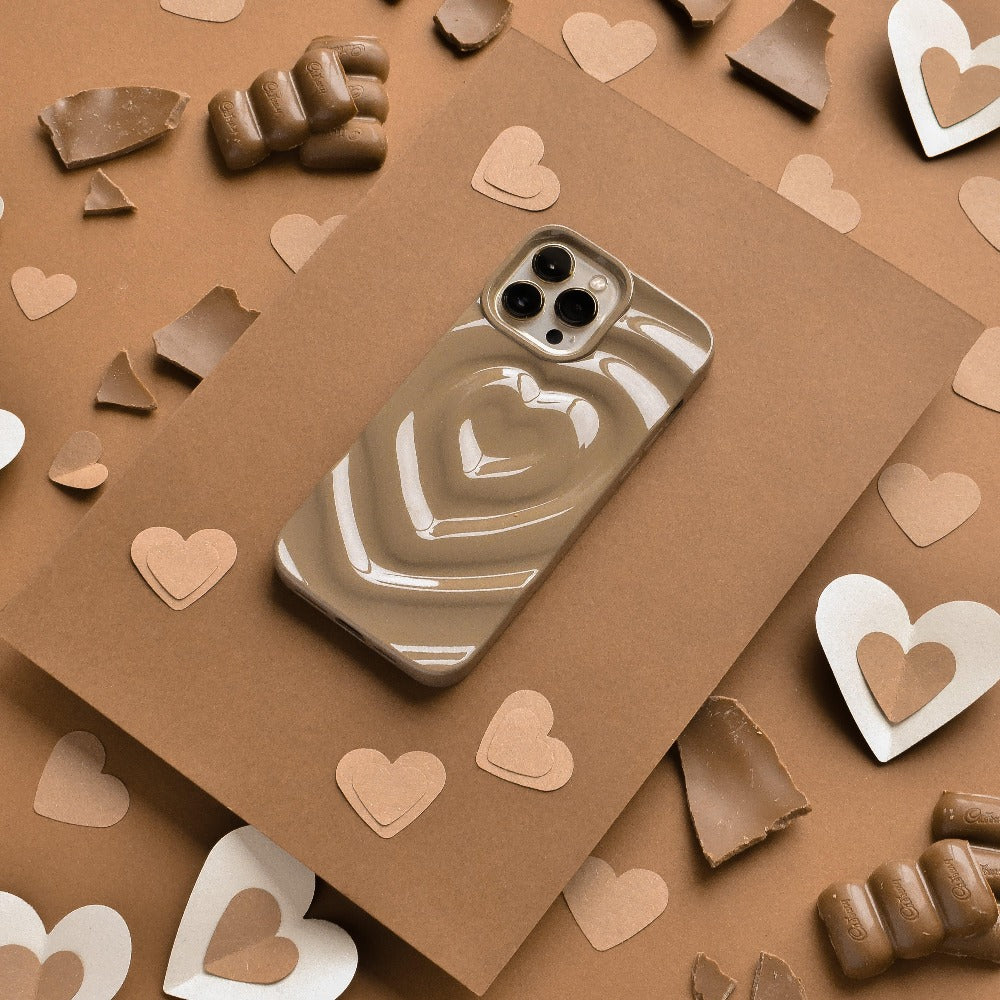 A smartphone adorned with the Coconut Lane Melting Heart Phone Case in Chocolate Brown is positioned on a brown rectangular board, encircled by various pieces of milk and white chocolate, including some heart-shaped ones. The background features a matching brown surface, enhancing the monochromatic aesthetic.