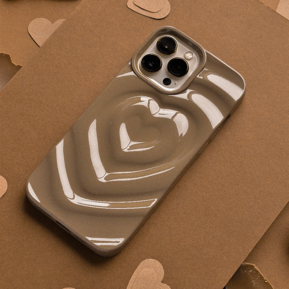A smartphone with a triple-lens camera is nestled in Coconut Lane's glossy, chocolate brown Melting Heart Phone Case, which features a raised heart design on the back. Resting on a taupe-colored cardboard surface, small heart cutouts are scattered around for added charm.
