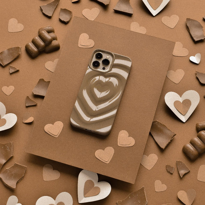 A smartphone with a Coconut Lane Melting Heart Phone Case - Chocolate Brown, featuring a brown and white heart pattern, rests on a chocolate brown surface surrounded by scattered heart-shaped paper pieces and chocolate chunks. The arrangement creates a warm, romantic aesthetic.