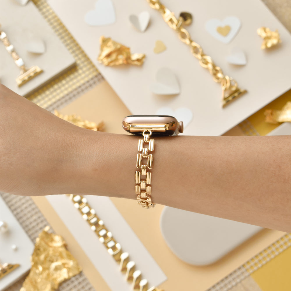 A close-up of a wrist adorned with the sophisticated Square Gold Bracelet Apple Watch Strap by Coconut Lane. The background showcases an array of gold jewelry pieces and charming heart-shaped cutouts, enhancing the luxurious and romantic atmosphere. The lighting accentuates the strap's luster and intricate craftsmanship.
