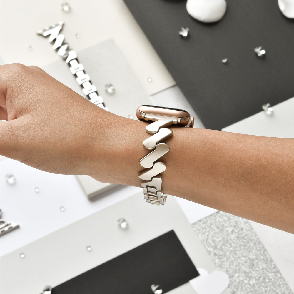 A hand is shown wearing an Apple Watch with the stylish Get Wavy Apple Watch Strap - Starlight by Coconut Lane. The background features various textures and colors, including black, white, and gray surfaces, along with scattered small metallic decorations. This stainless steel adjustable strap fits all Apple Watch models and enhances the modern, minimalist aesthetic.