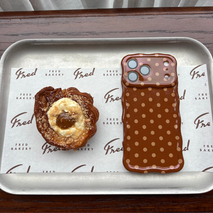 Flat lay photo in a cafe of the Dot Society Wavy Chocolate Brown polka dot case. The case is chocolate brown with lighter brown polka dots. Next to it is a bakery pastry on the tray