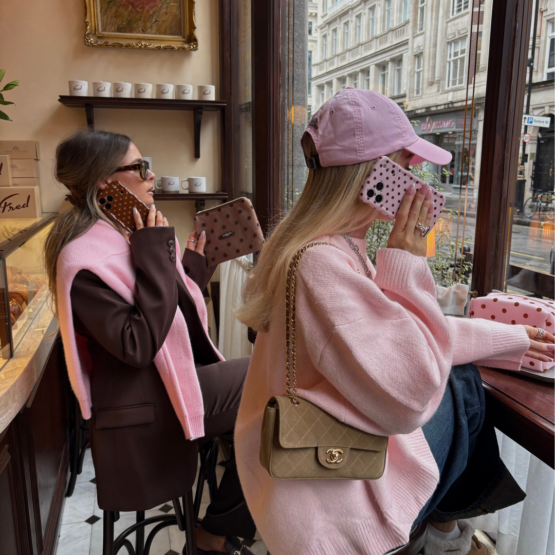 Two woman in a cafe wearing baby pink and brown outfits, both holding an iphone up to their ear. The iphones have a wavy baby pink phone case with brown polka dots and a wavy brown with lighter brown polka dots. 