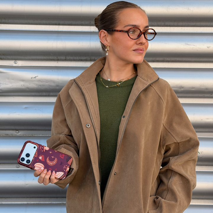 A woman with glasses and her hair in a bun holds a phone with a decorative  autumn-themed case featuring illustrations of pumpkins, a crescent moon, and a slice of pumpkin pie.