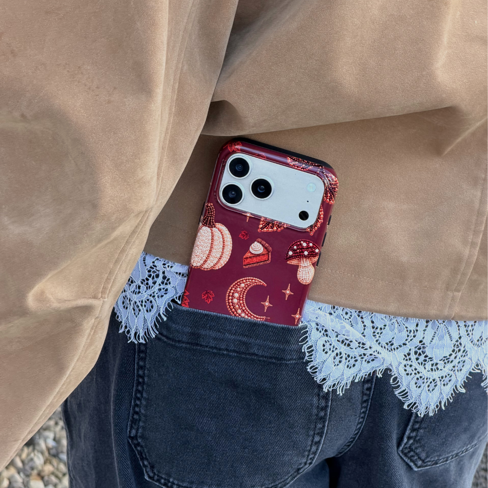 A close-up lifestyle shot of a phone case tucked into the back pocket of a person's jeans. The person is wearing a tan jacket with white lace trim and dark blue jeans. The phone is in a decorative  autumn-themed case featuring illustrations of pumpkins, a crescent moon, and a slice of pumpkin pie.
