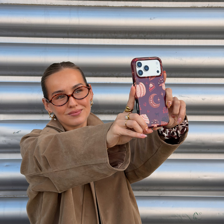 A woman with glasses and her hair in a bun holds a phone with a decorative  autumn-themed case featuring illustrations of pumpkins, a crescent moon, and a slice of pumpkin pie.