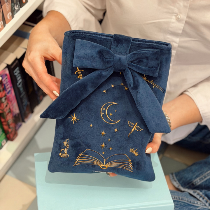 Book Sleeve made from soft navy blue velvet with gold embroidered crescent moon, stars, crown and open book illustrations, finished with a large velvet bow and Coconut Lane logo.