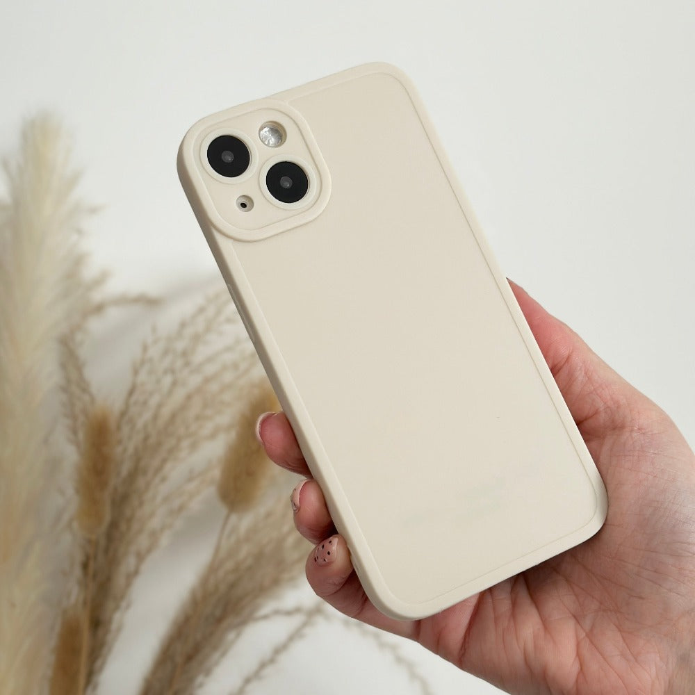 A hand holding a light beige smartphone adorned with the NAKD Phone Case in Creamy White by Coconut Lane, featuring a minimalistic design. The phone has a dual camera setup and a flash on the top left corner of the back. The background showcases out-of-focus beige and white pampas grass against a plain white backdrop.