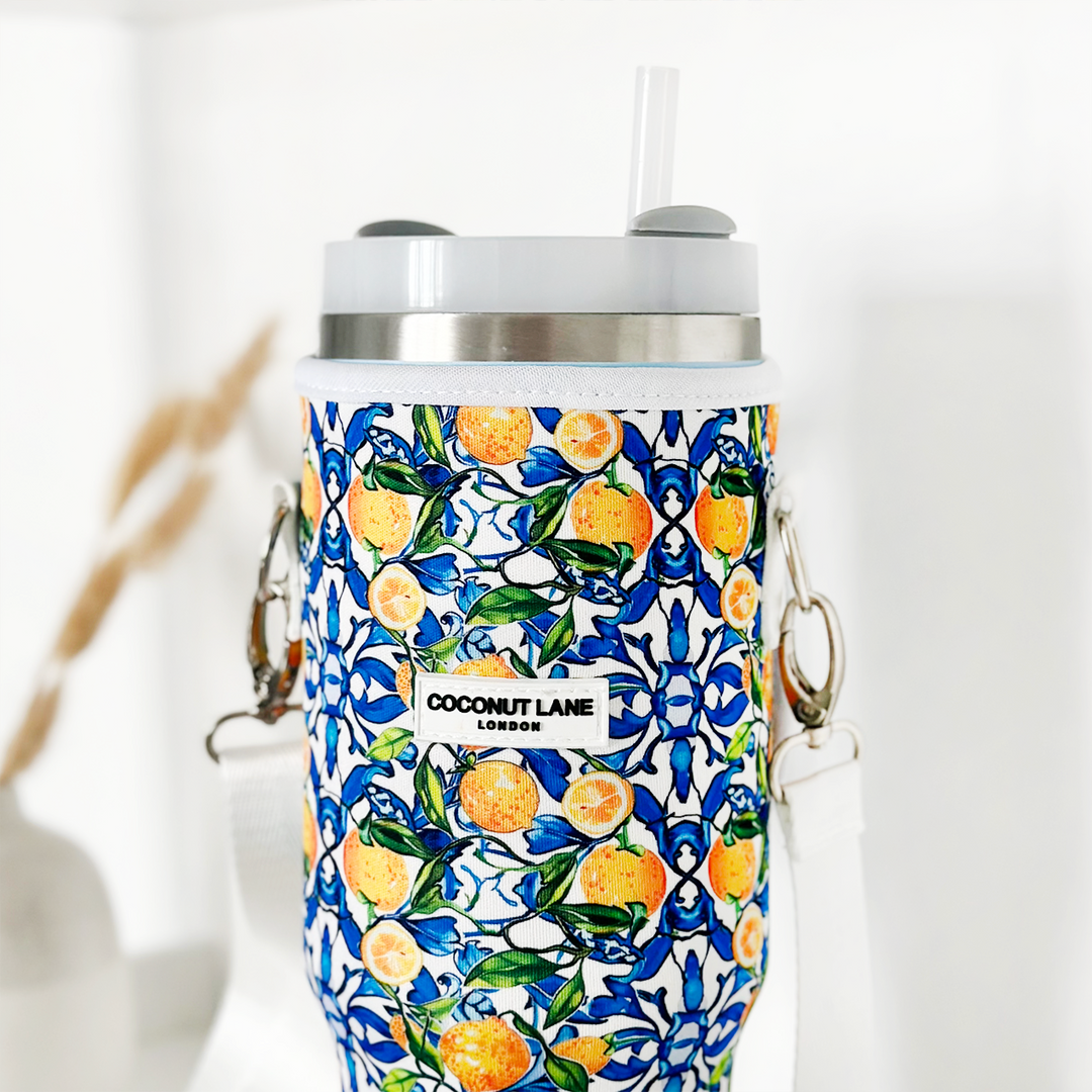 The Tumbler Carry Case - Amalfi Lemon by Coconut Lane is a reusable travel mug adorned with a vibrant sleeve featuring lemons and green leaves on a blue and white background. It comes with a grey lid, clear straw, and detachable white strap. The sleeve is printed with "Coconut Lane London," making it perfect for stylish hydration on the go.