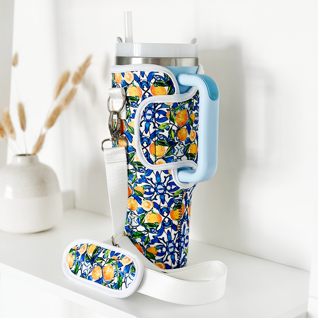 A tall, reusable Stanley Quencher travel mug with a straw, featuring a blue floral and lemon patterned sleeve and a light blue handle, stands on a white surface. Next to it is Coconut Lane's Tumbler Carry Case - Amalfi Lemon, complete with a shoulder pad. In the background, there is a minimalist vase holding dried pampas grass.