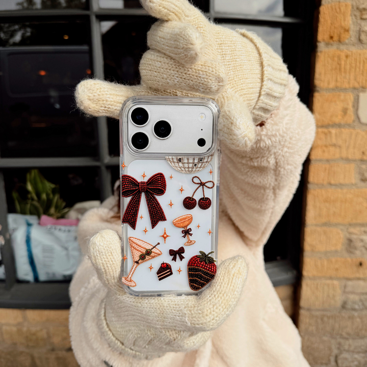A woman wearing cream knit gloves and a cream teddy coat, holds up a clear phone case decorated with a red bow, cherries, a martini glass, a strawberry dipped in chocolate, and small star accents. The background shows a cafe setting.