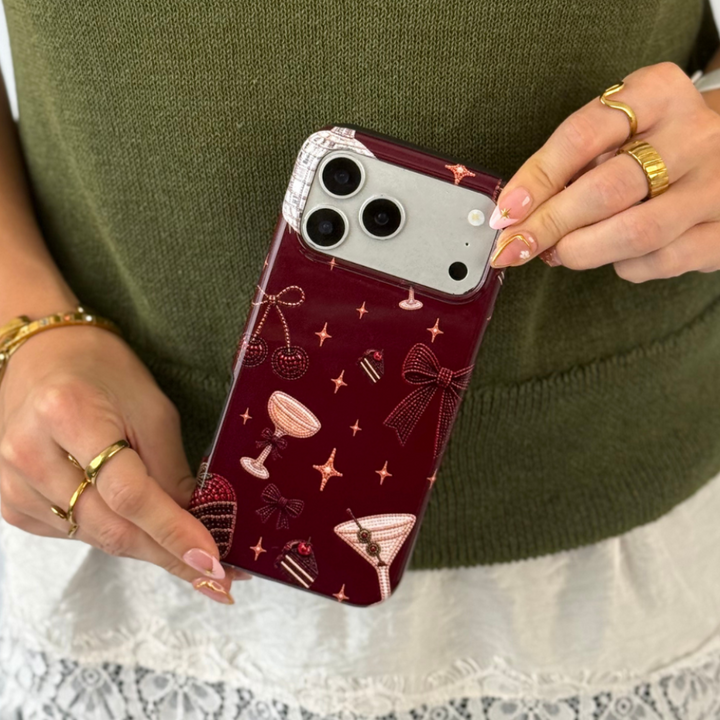 Woman in a dark green jumper with a white lace shirt underneath holds a burgundy phone case with decorative beaded illustrations on it, such as a bow, a strawberry, a cocktail, stars and cherries. 