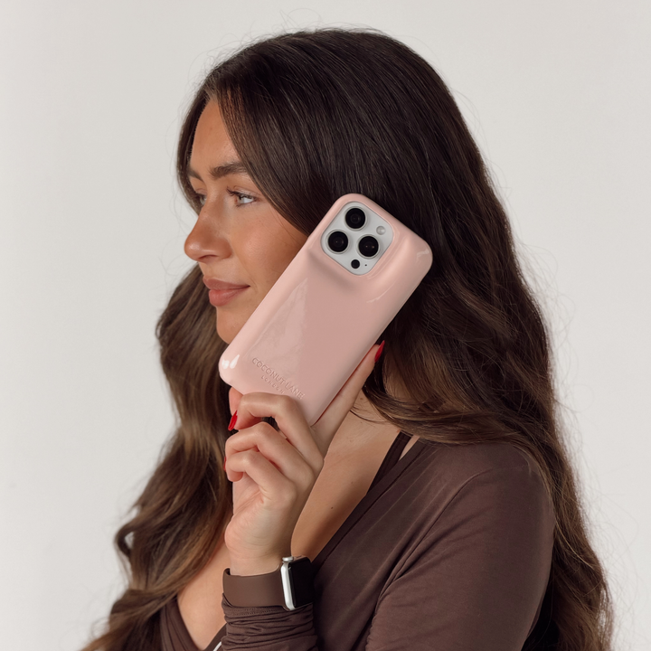 Woman in a brown pilates outfit is holding an iphone up to her ear. The iphone is in a glossy pastel pink Coconut Lane puffer phone case with embossed logo. On her wrist is a Coconut Lane NAKD Chocolate brown apple watch strap.