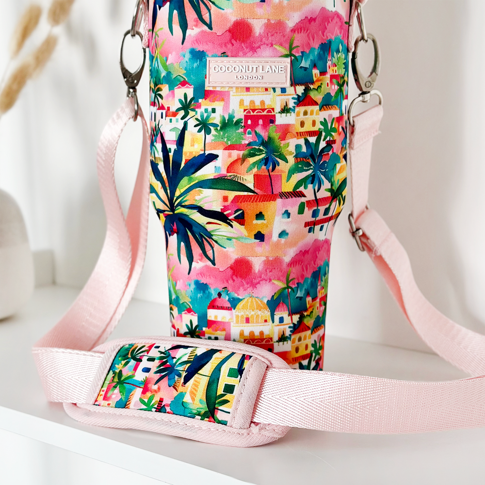 The Tumbler Carry Case - Portofino Dreams by Coconut Lane is a colorful, tropical-themed accessory adorned with vibrant illustrations of palm trees and houses. It features a pink fabric strap, silver metal clasp, and a tag that reads "Coconut Lane London." The holder is positioned on a white surface.
