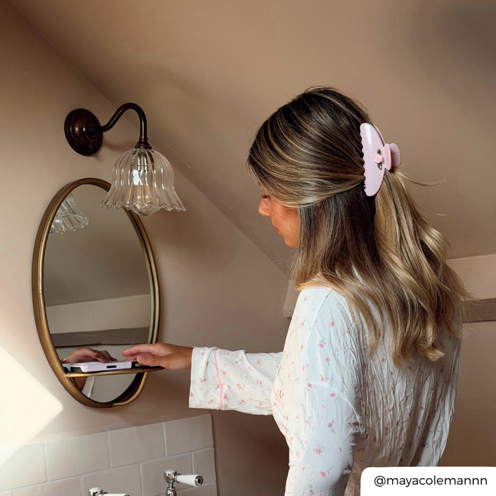 A woman with blonde-highlighted hair wearing a white long-sleeve top with small pink floral prints and a light pink claw clip, seen from behind as she looks into a bathroom mirror. 