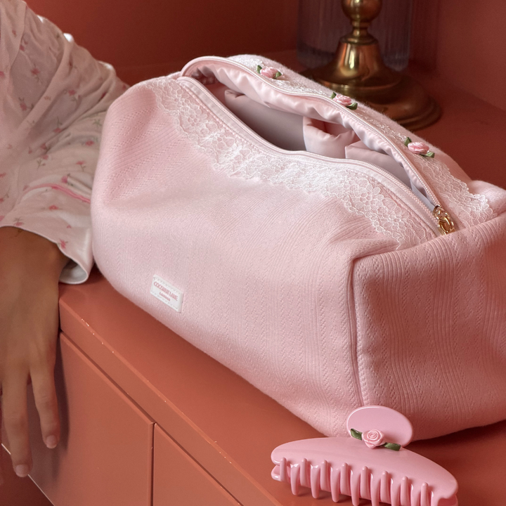 A pink hair tool bag with white lace trim and a gold zipper, resting on a peach-colored cabinet next to a person's hand. A matching pink hair claw clip is in the foreground.