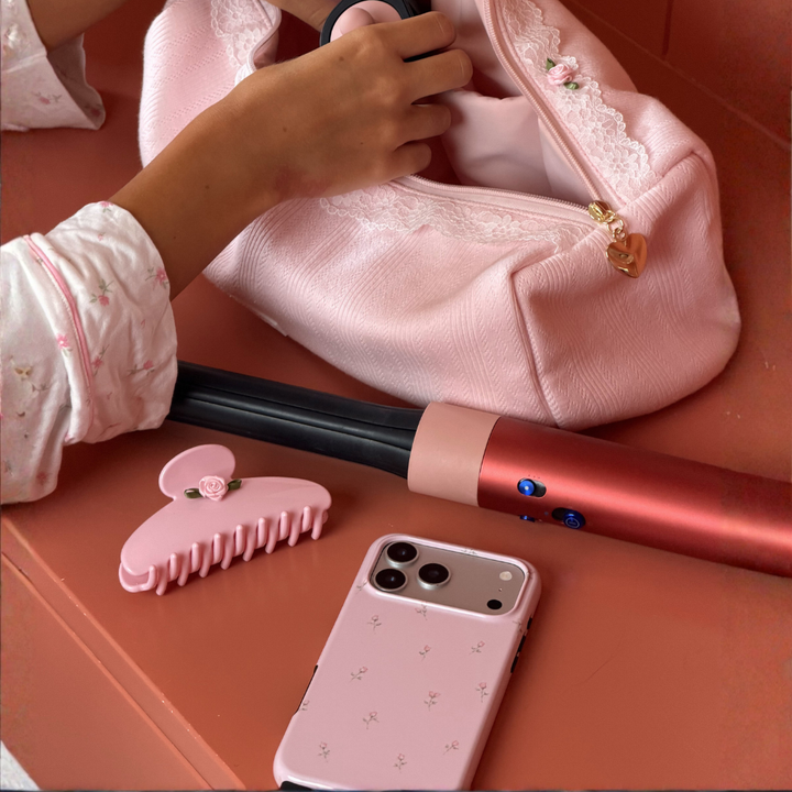 A pink fabric hair tool bag with lace trim, with a person's hand reaching into the bag. A pink hair wand, a pink claw clip, and a pink phone case are also arranged nearby. The phone case has dainty floral pattern.