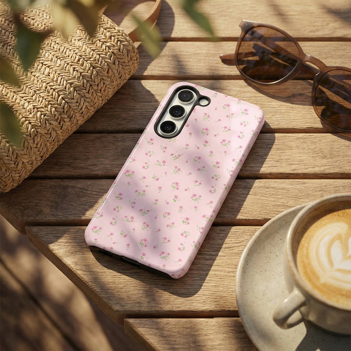 Pink Samsung phone case with ditsy floral pattern on table next to sunglasses, woven bag, and a cup of coffee.