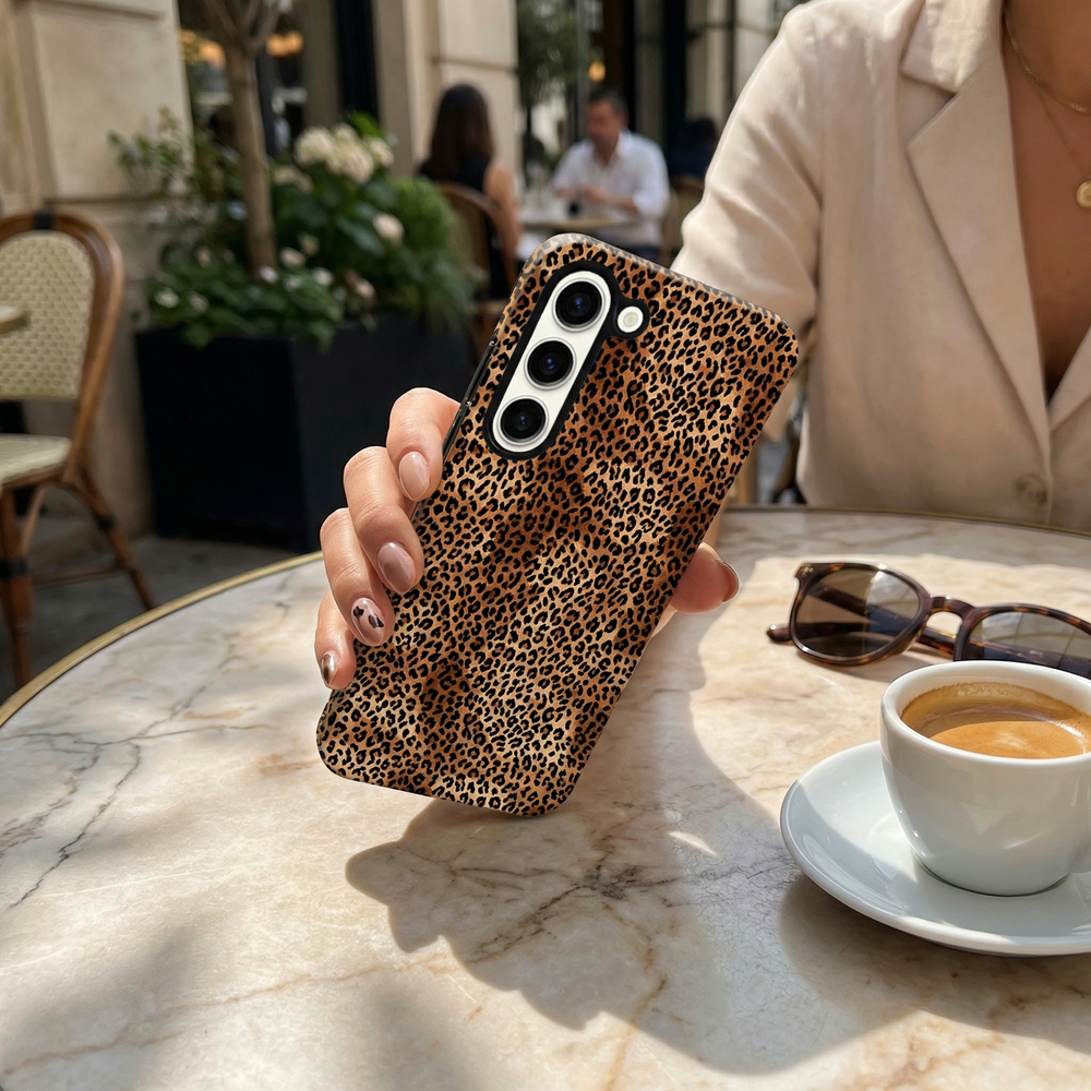 Hand holding a Coconut Lane Samsung tough phone case with leopard print at an outdoor café table with coffee and sunglasses.