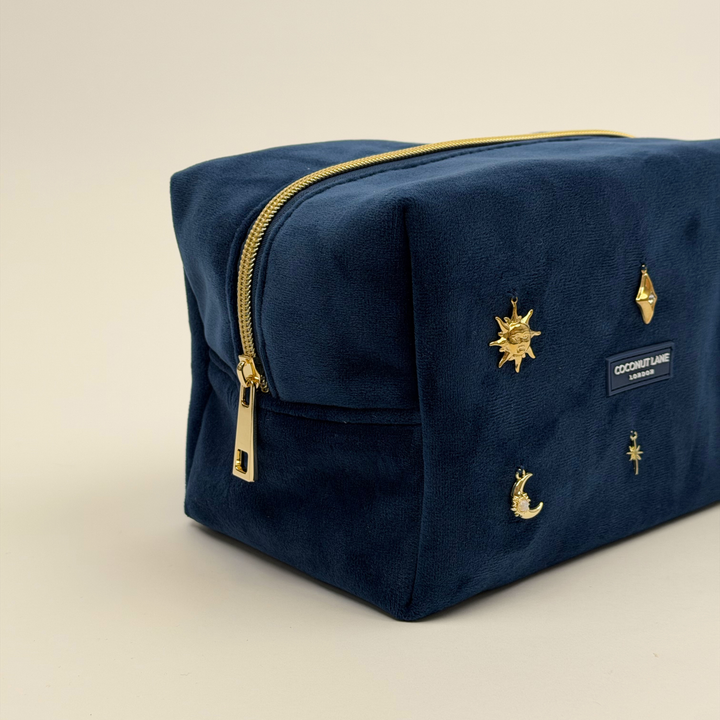 Stardust After Dark makeup bag. Dark navy blue velvet material with gold zip, and sewn-on gold celestial-themed charms including a sun, moon, star, diamond shape and 11:11 charm.  