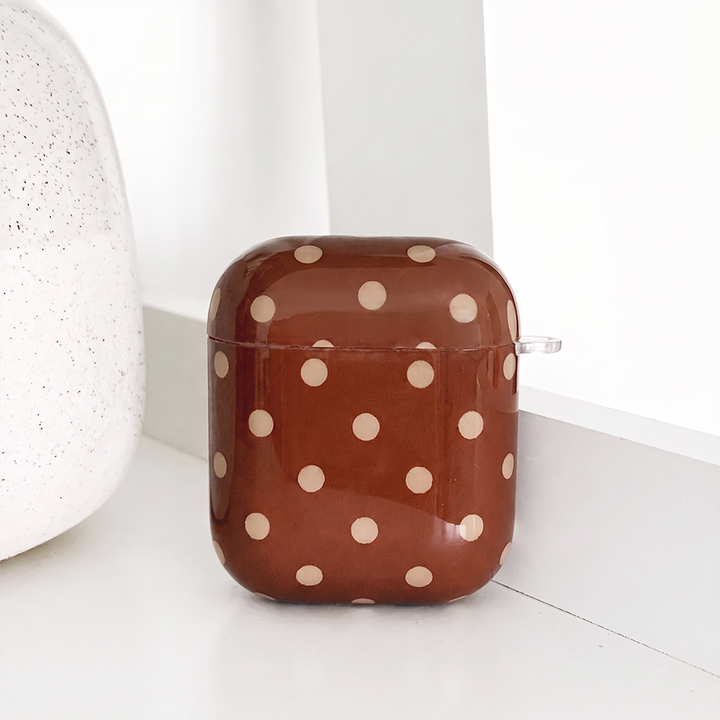 A glossy brown case for AirPods with cream polka dots and a small clear clip loop on the side. It sits on a white shelf beside a white speckled vase.