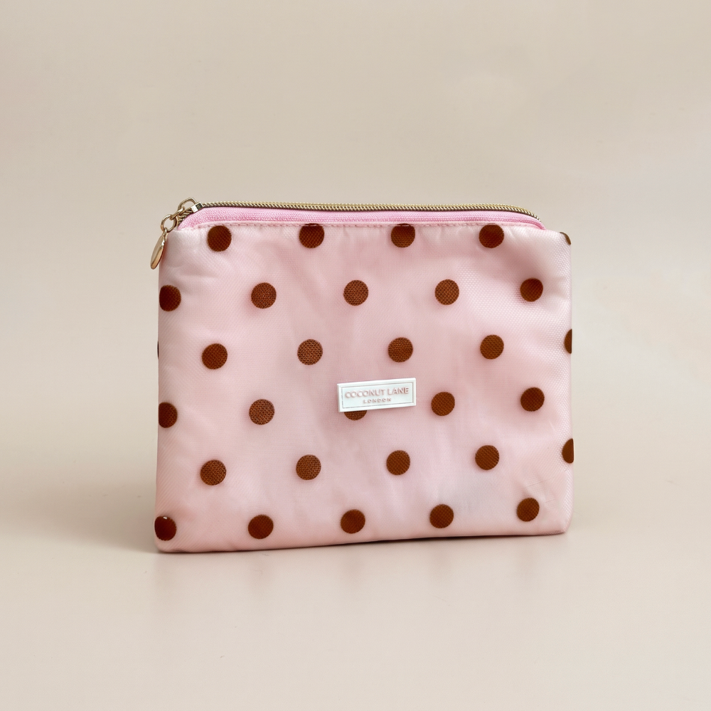 Pink mesh pouch with brown polka dots, a gold top zip with a heart-shaped zip pull, and a white Coconut Lane London logo badge on the front, shown on a beige background.