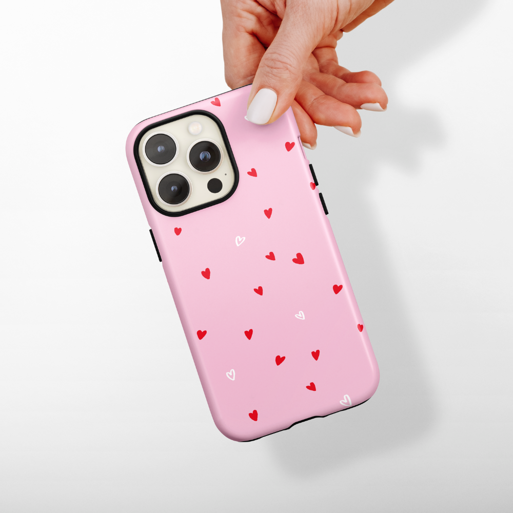 A hand with white nail polish is holding an iPhone adorned with Coconut Lane's Tough Phone Case - Cute Hearts, featuring small red and white hearts against a pink backdrop. The setting is a plain white background.