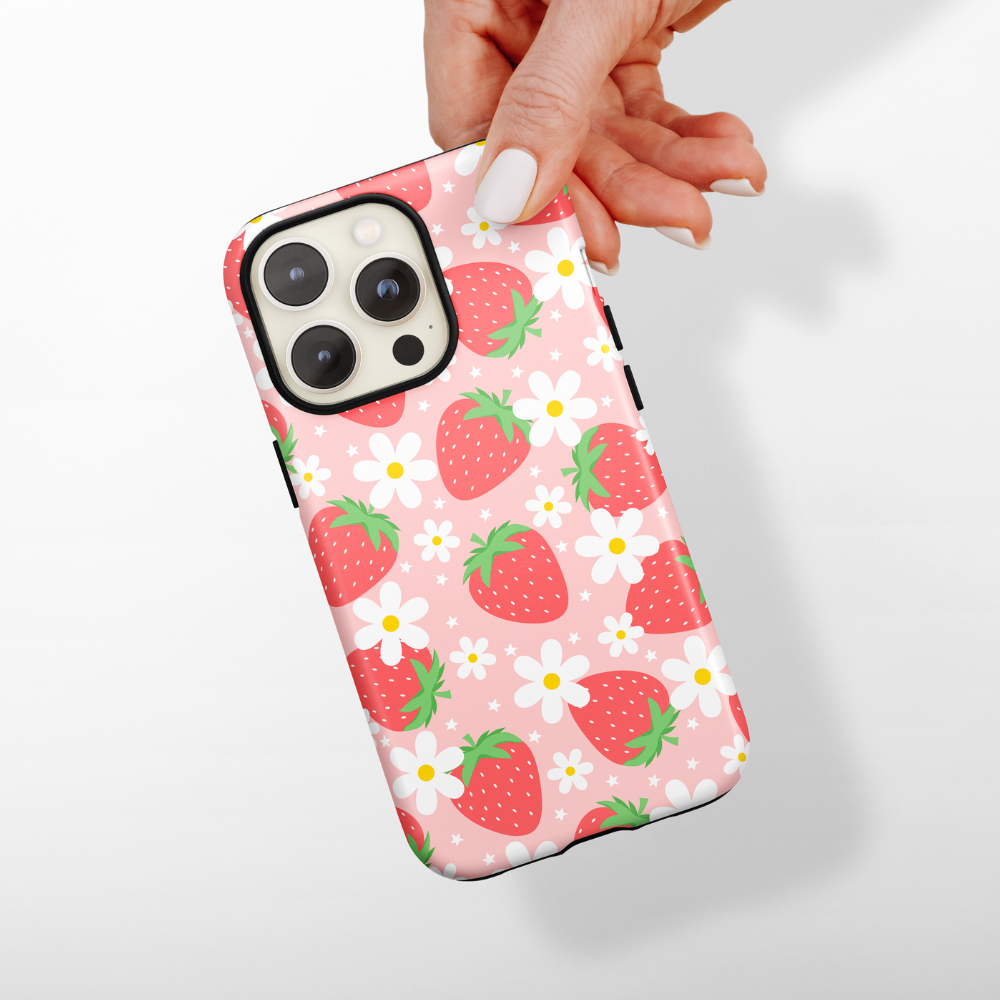 A hand with white-painted nails holds an iPhone encased in Coconut Lane's Tough Phone Case - Berry Cute Strawberry. The pink, protective case is adorned with red strawberries and white flowers, showcasing a cute and playful design against a neutral, blurred background.