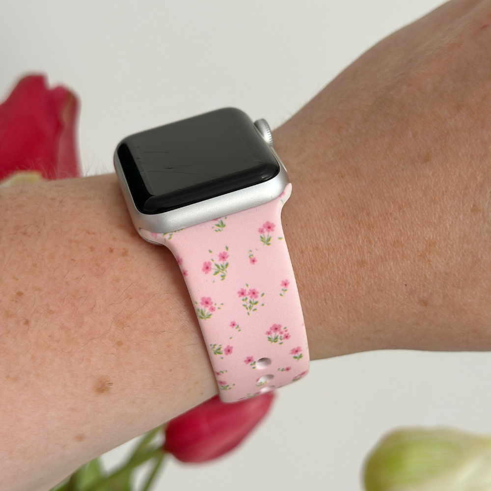 Pink floral Apple Watch strap by Coconut Lane, worn on wrist with red and white flowers blurred in the background.