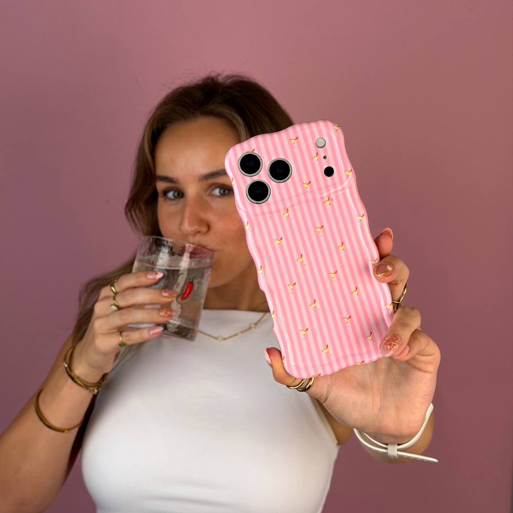 A woman taking a selfie, holding a wavy pink striped phone case with small spicy margarita illustrations, held by a person wearing a white top and black jeans against a solid pink background. She is sipping from a glass with red chillis on it. 