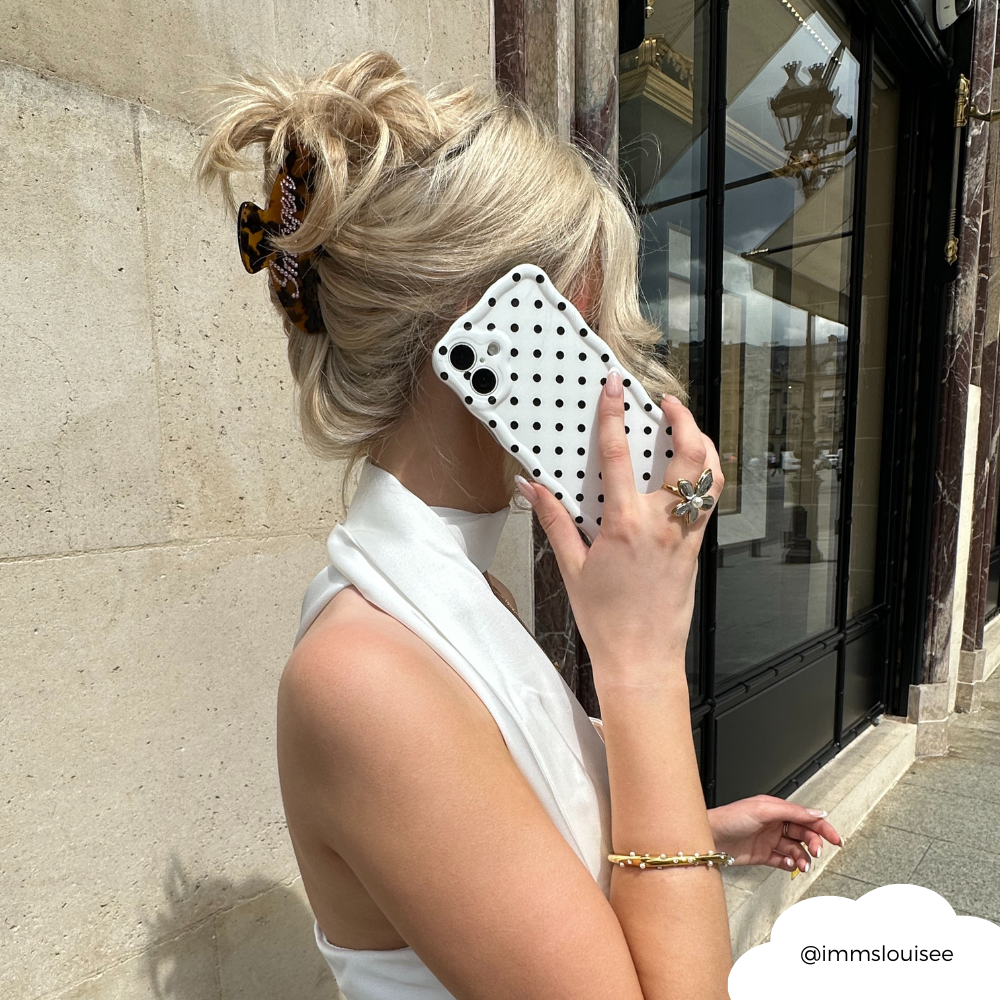 Blonde woman with hair clip holds a white polka dot wavy phone case to her ear outside a building.