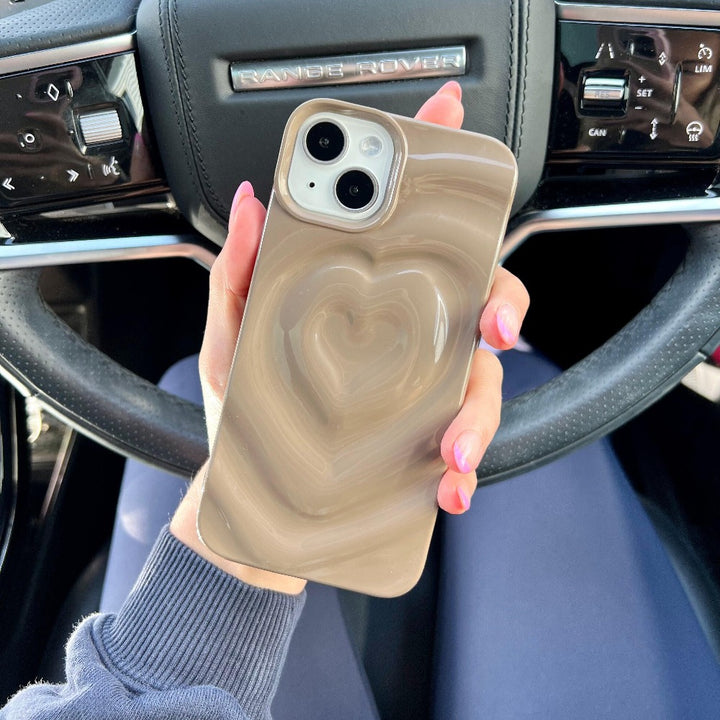 A hand with pink nail polish holds a Coconut Lane Melting Heart Phone Case in chocolate brown, showcasing its 3D melting heart effect. The person is seated in the driver's seat of a luxury vehicle, with a visible steering wheel and dashboard controls in the background.