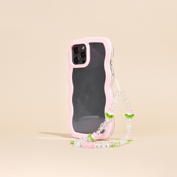 A smartphone case with a wavy pink edge, protecting a phone with triple rear cameras, is shown standing upright. Attached to the case is the Beaded Phone Strap - Cute Tulips by Coconut Lane, featuring small green leaves and pink beads that resemble cherries. This charming phone accessory stands against a plain beige background.