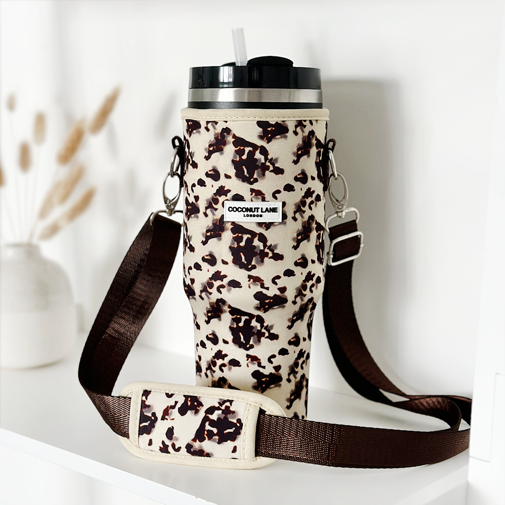 A tall, reusable Stanley Quencher travel mug with a black lid and straw is fitted into a stylish Ivory Tort Tumbler Carry Case holder with a brown strap and shoulder pad. The holder has a label that reads "Coconut Lane." The background is minimal, featuring a white surface and a decorative vase with dried plants.