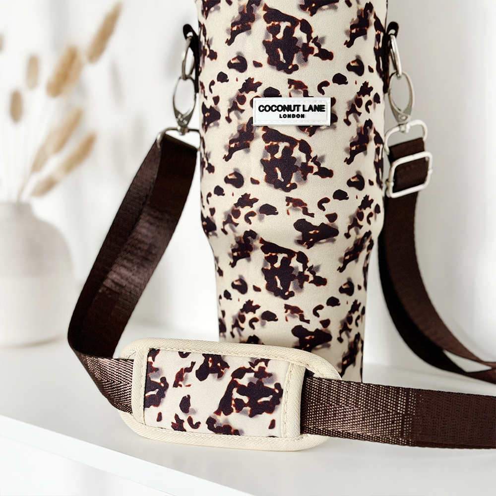 A close-up of the stylish Tumbler Carry Case - Ivory Tort, featuring an elegant animal-print design and a brown adjustable strap, perfect for carrying your travel mug. The brand label on the case reads "Coconut Lane." In the background, a blurred white vase with dried pampas grass is visible on a white surface.