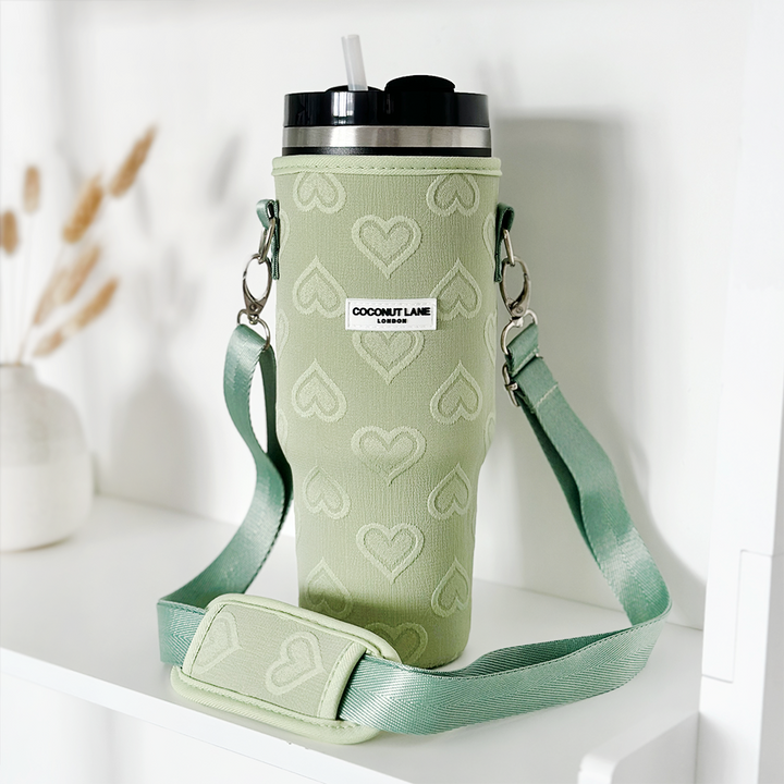 A green insulated Stanley Quencher tumbler, featuring a black lid and white straw, is showcased alongside a matching "Tumbler Carry Case - Sage Heart." The travel mug bag from Coconut Lane is decorated with heart patterns and includes a shoulder strap with the "Coconut Lane" label. In the background, dried plants and a white vase are visible.