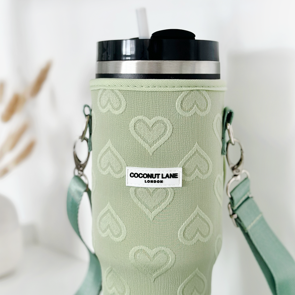 A Stanley Quencher tumbler adorned with a Sage Heart Tumbler Carry Case from Coconut Lane, featuring a green heart-patterned sleeve. It has a silver and black lid with a straw and includes a green strap with metal clasps. The sleeve is labeled "Coconut Lane London," set against a blurred background of white decor elements.