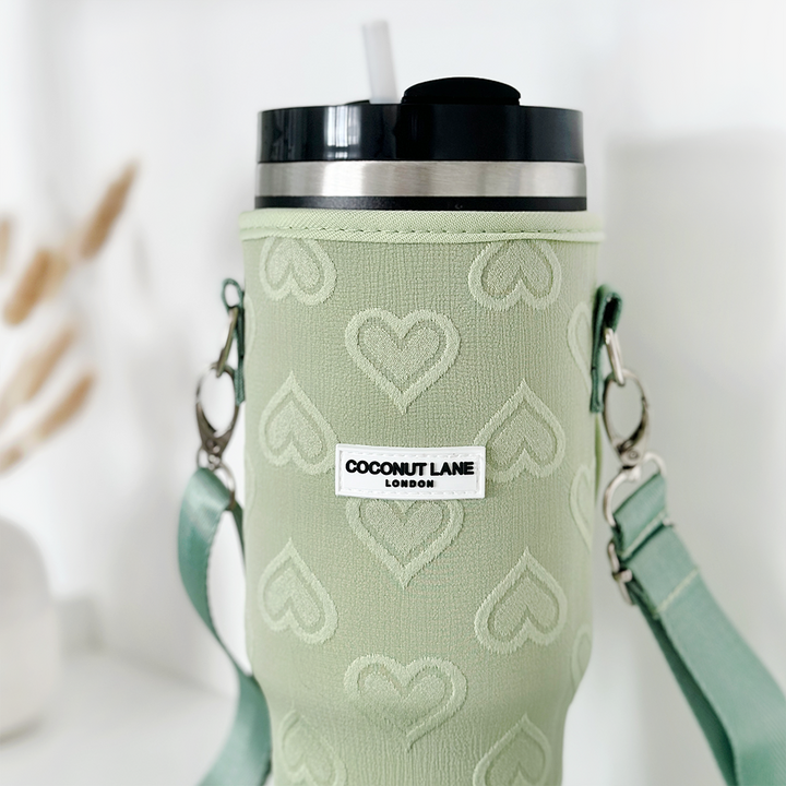 A Stanley Quencher tumbler adorned with a Sage Heart Tumbler Carry Case from Coconut Lane, featuring a green heart-patterned sleeve. It has a silver and black lid with a straw and includes a green strap with metal clasps. The sleeve is labeled "Coconut Lane London," set against a blurred background of white decor elements.