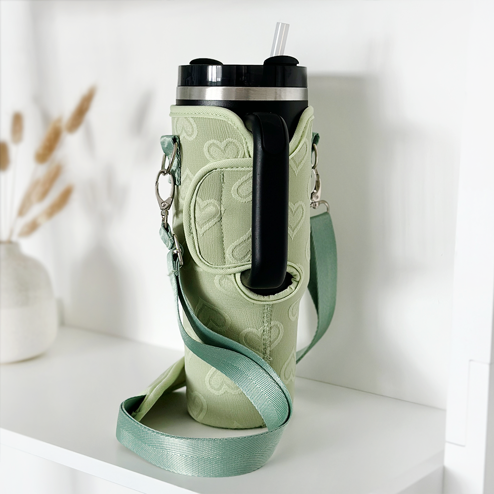 A light green Stanley Quencher with a black lid and transparent straw sits in a Tumbler Carry Case - Sage Heart by Coconut Lane, placed on a white shelf. The carry case has a matching green color with a subtle heart pattern and metal clips. In the background, there is a white vase with dried grass.