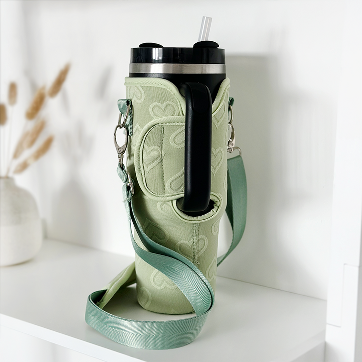 A light green Stanley Quencher with a black lid and transparent straw sits in a Tumbler Carry Case - Sage Heart by Coconut Lane, placed on a white shelf. The carry case has a matching green color with a subtle heart pattern and metal clips. In the background, there is a white vase with dried grass.
