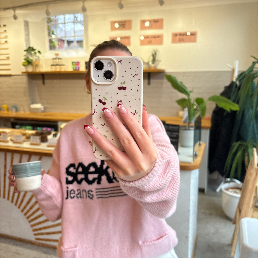 Pink sweater, face hidden by Coconut Lane phone case with a ditsy bows and cherries pattern in a café mirror selfie.