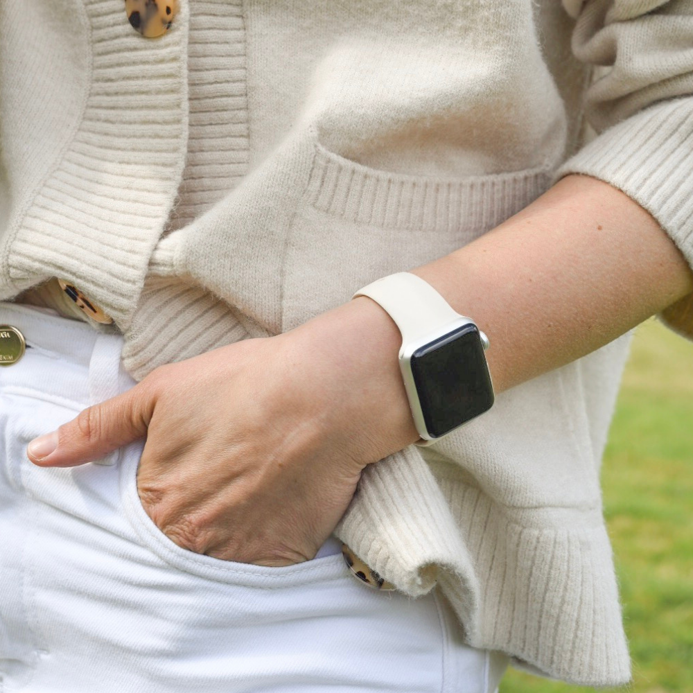 A person wearing a beige cardigan and white pants has their left hand in their pants pocket. They are also sporting an Apple Watch with the NAKD Apple Watch Strap - Almond White by Coconut Lane on their wrist. The background is a grassy outdoor area.