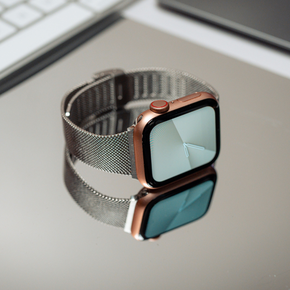 A smartwatch featuring the Coconut Lane Milanese Apple Watch Strap - Silver lies on a reflective surface. The watch face displays an analog clock with simple white hands. Positioned near a keyboard and a laptop, with their edges partially visible in the background, this strap is compatible with Apple Watch models.