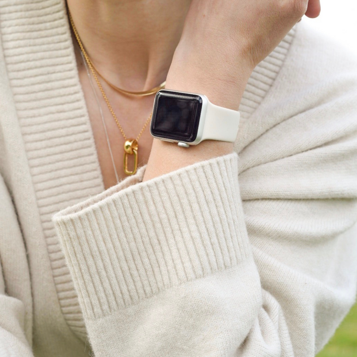 A person wearing a cream-colored sweater is shown with an Apple Watch featuring the NAKD Apple Watch Strap - Almond White from Coconut Lane on their wrist, along with two gold necklaces, one adorned with a geometric pendant, around their neck. The camera angle highlights their arm and chest, with the watch face turned sideways.