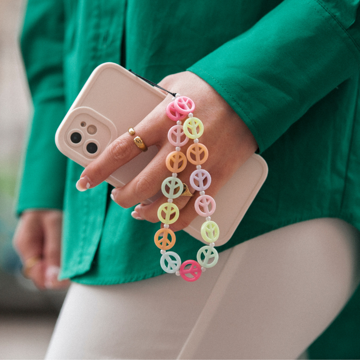 A person wearing a green shirt is holding a white smartphone with a colorful peace sign wrist strap. The phone, encased in the NAKD Phone Case - Creamy White by Coconut Lane, is secured by the strap looped around their fingers. The background is blurred, highlighting the hand and phone.
