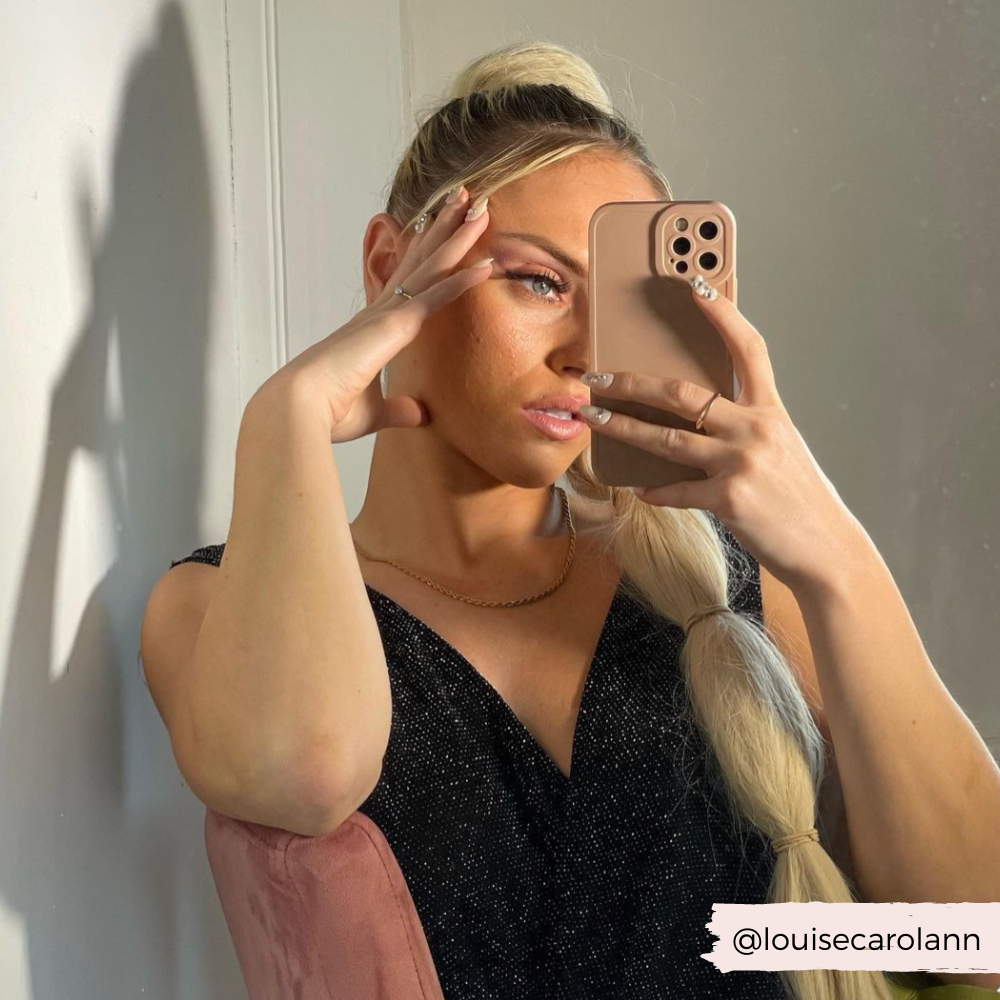 
A person with long blonde hair in a ponytail takes a mirror selfie using a phone encased in an elegant NAKD Phone Case - Nude from Coconut Lane. They wear a black sleeveless top and a gold necklace, holding their head with one hand. The background is neutral and the Instagram handle "@louisecarolann" is visible.